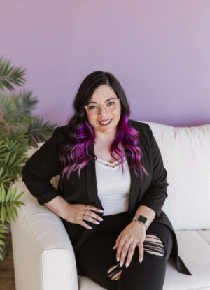 A woman with long black and purple hair, glasses, and a black blazer sits on a white couch against a light purple wall, smiling at the camera—ready to start a 1:1 Mentoring Session.