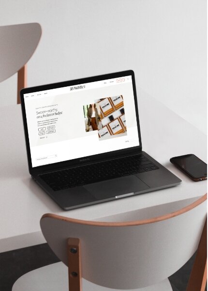 A black laptop displaying a website sits on a white table next to a smartphone, with two white chairs nearby—perfect for discussing eCommerce platform comparison or exploring website builder options like Showit vs Shopify.