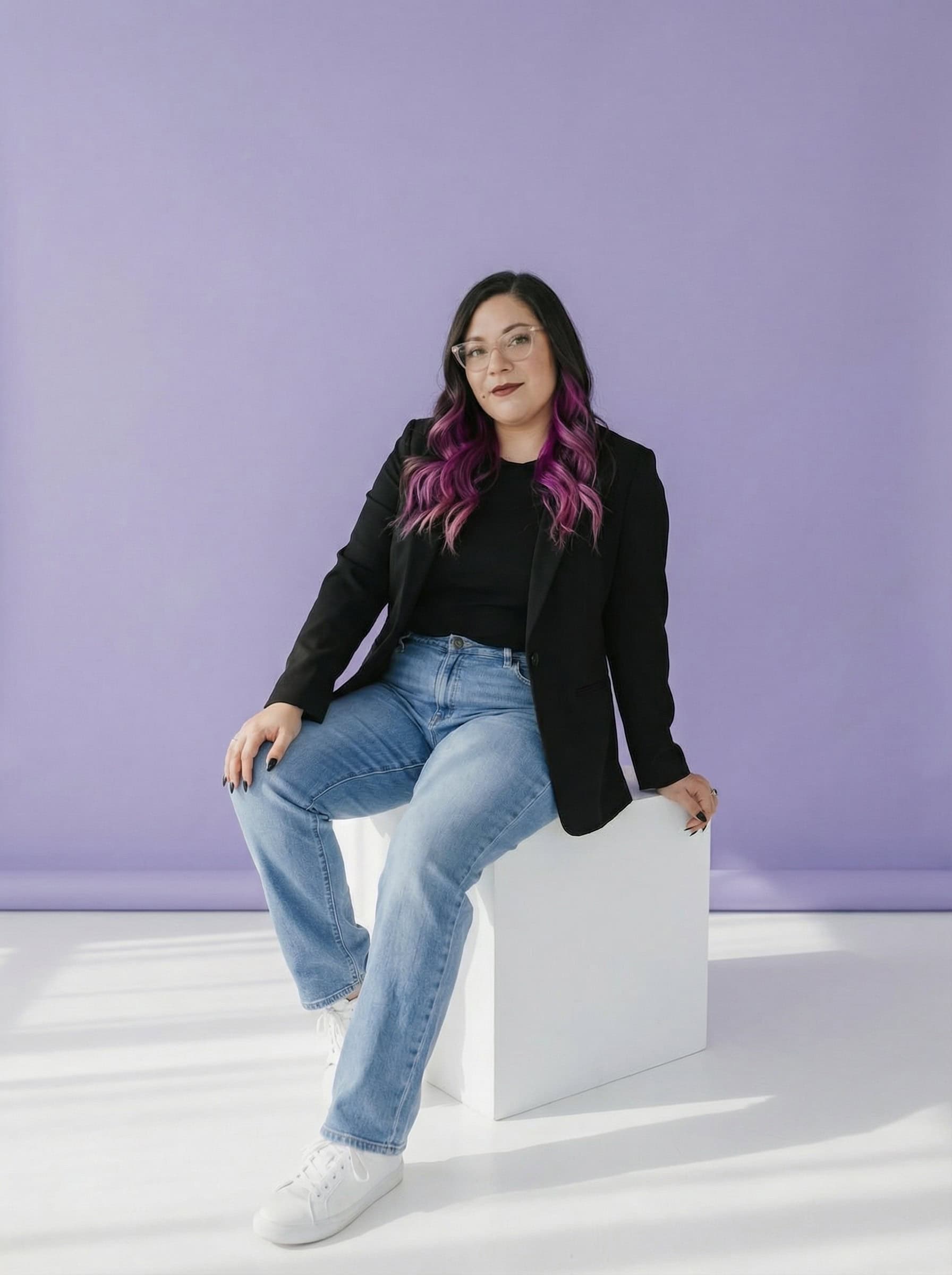 Ingrid Urena with long purple hair, glasses, and casual clothes sits on a white cube against a light purple background.