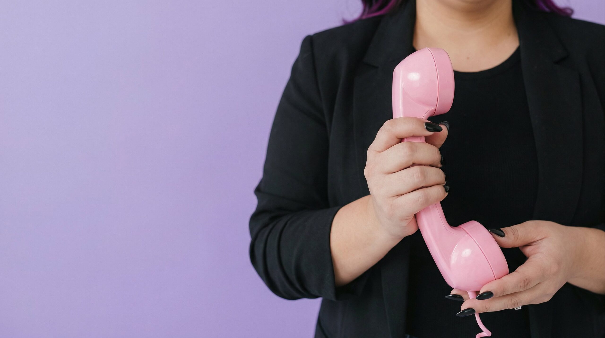 A person in a black blazer holds a pink telephone handset against a plain purple background.